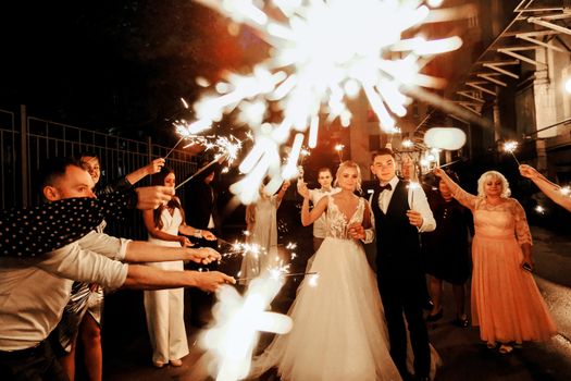Happy young bride and groom standing on street near unrecognizable guests with burning sparklers during wedding party in evening