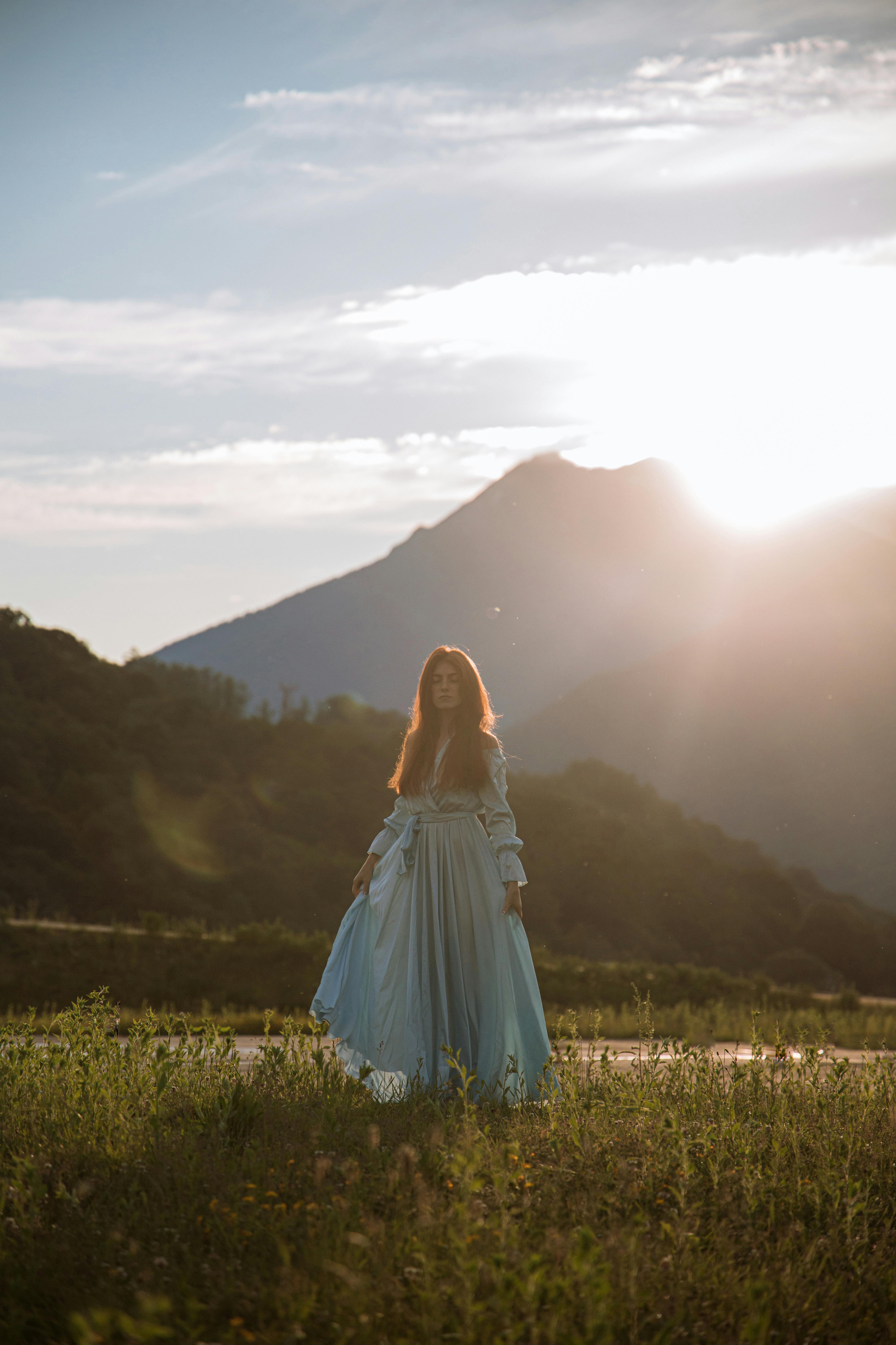 一位身着蓝色连衣裙的女子，站在田野里，身后是山脉和日落的天空