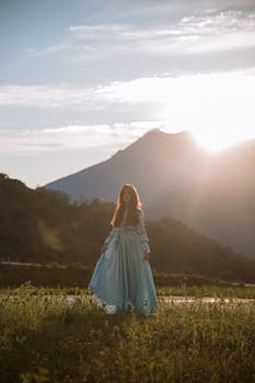 一位身着蓝色连衣裙的女子，站在田野里，身后是山脉和日落的天空