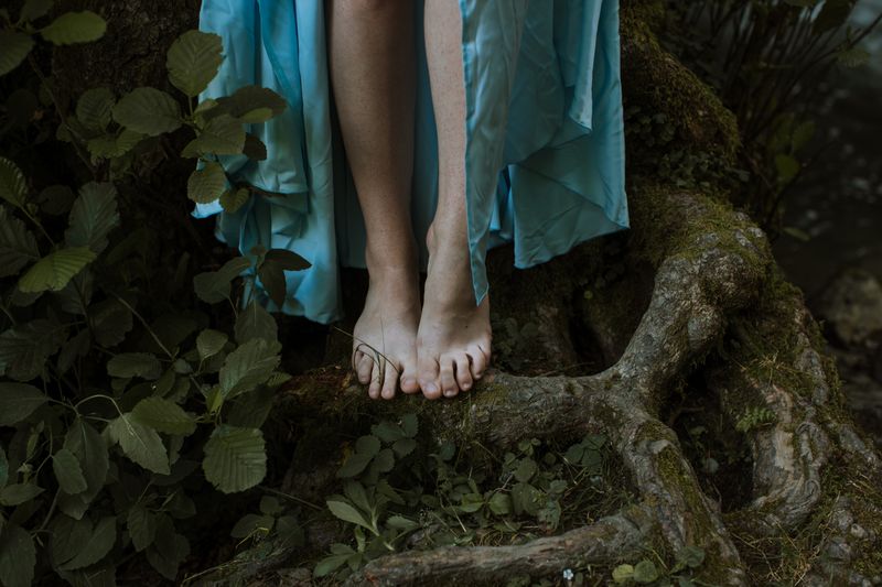 Close-up of bare feet standing on textured tree roots surrounded by green leaves