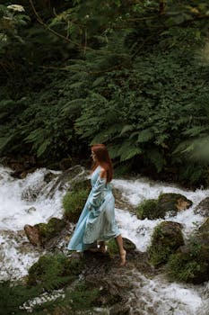 Red-haired woman in blue dress standing by a flowing creek in a lush forest setting.