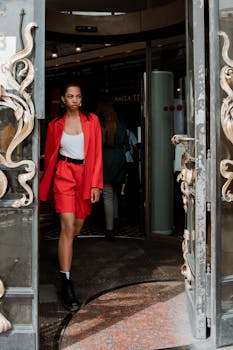 Stylish woman in red suit exits ornate doorway, capturing city elegance and modern fashion.
