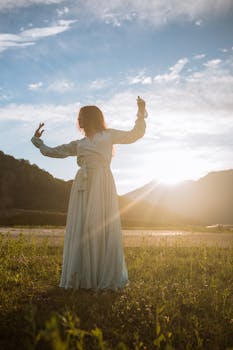 A woman in a flowing dress dancing gracefully in a sunlit meadow with mountains in the background.