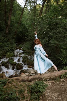 A woman in a blue dress gracefully dances on a log in a lush forest beside a flowing stream.
