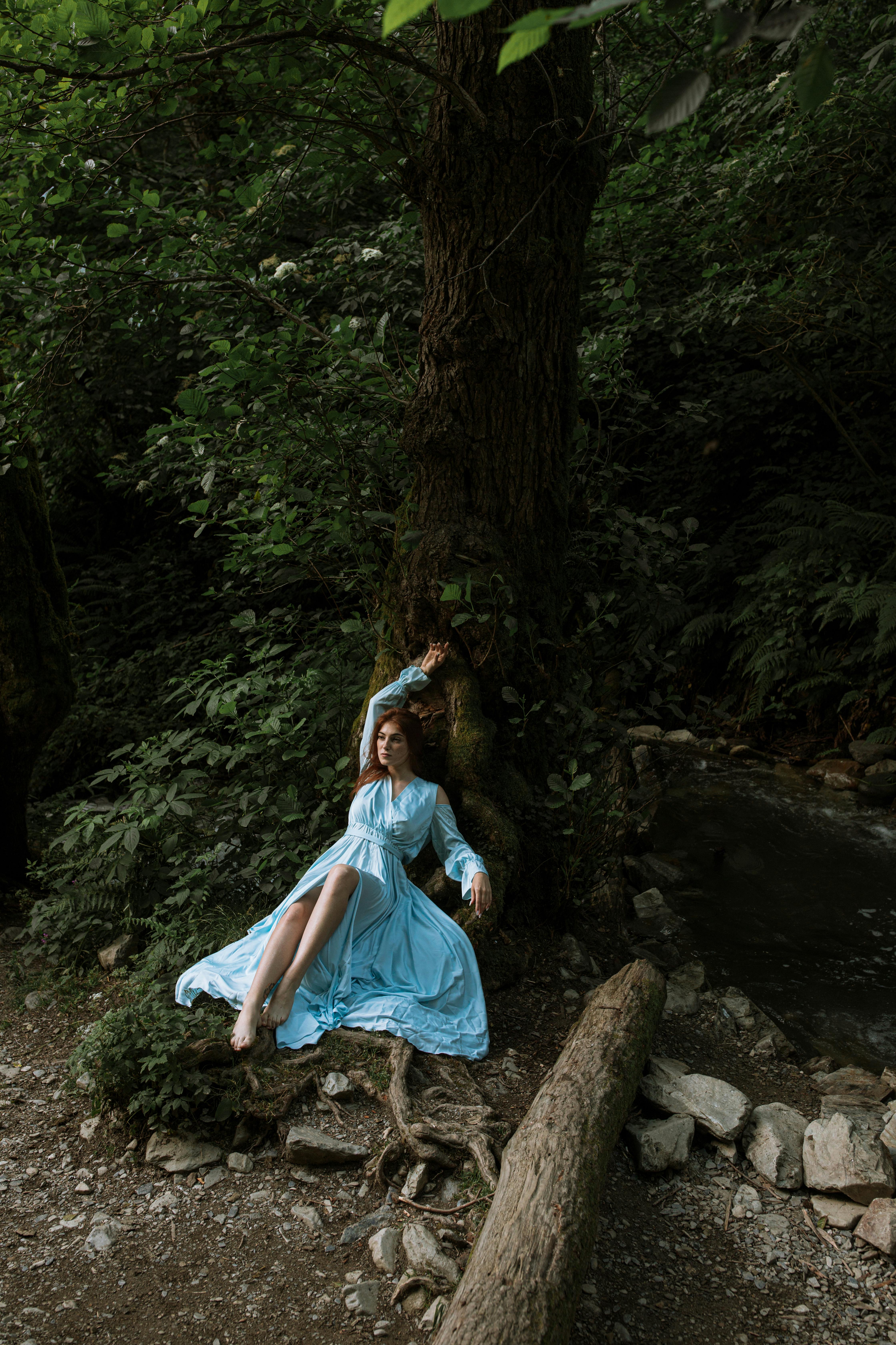 在郁郁葱葱的森林里，一位身着飘逸蓝色连衣裙的女子正倚着树干休息