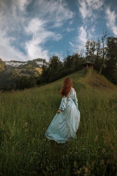 A red-haired woman in a blue dress walks gracefully through a lush green field during daytime.