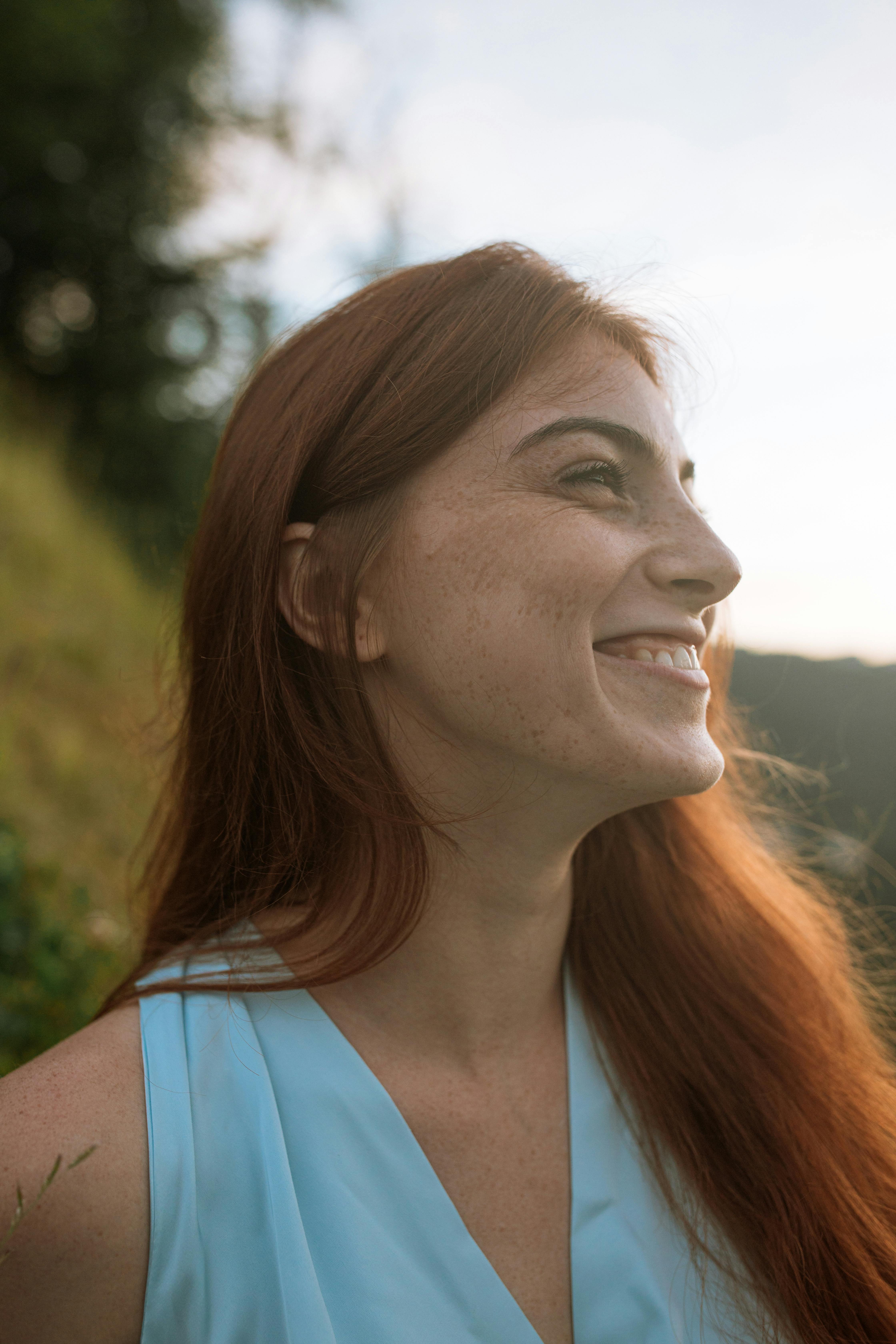 A close-up portrait of a smiling woman with red hair and freckles enjoying a sunny day outdoors.