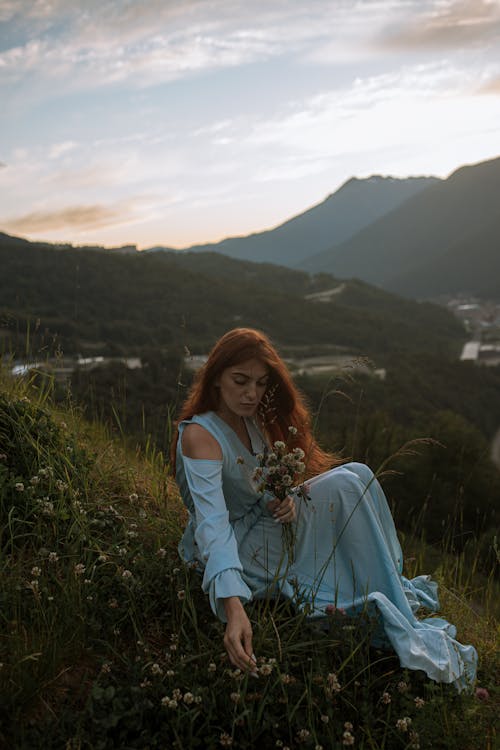 Photo of a Woman in a Blue Dress Picking Up Flowers