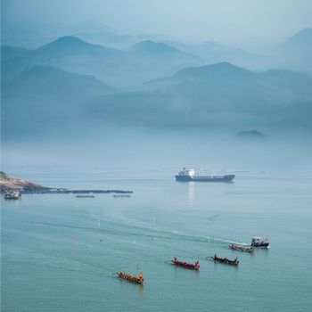 龙舟飞越雾蒙蒙的海面，远处可见货船与山脉