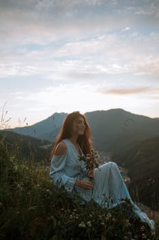A woman in a blue dress sits on a grassy hill, enjoying the mountain sunset.