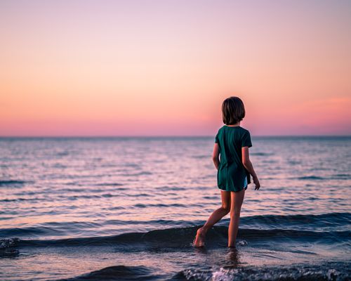 Free Back view of faceless little girl in casual clothes standing in sea with waves and enjoying sunset Stock Photo