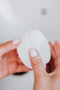 Close-up of a woman's manicured hands holding a white face cream jar against a blurred background.