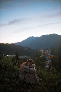 黄昏时分，一位穿着花裙的女人坐在草地上，四周环绕着山脉和树木