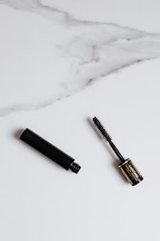 A minimalist image of a mascara brush and its container lying on a marble surface.
