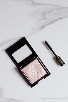 Flat lay of mascara and highlighter compact on a marble background.
