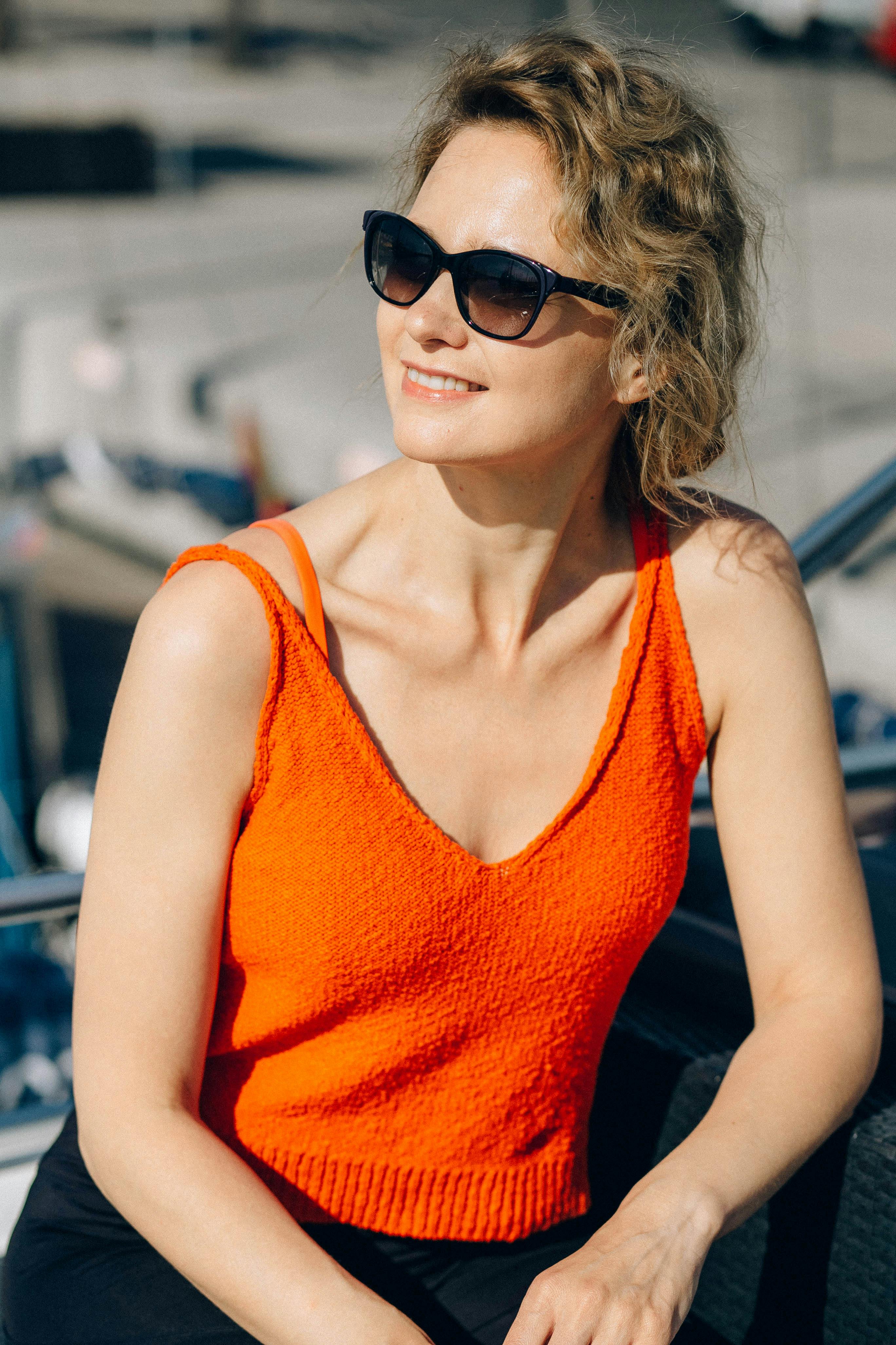 一位穿着橙色上衣、戴着太阳镜的女人在户外灿烂地微笑，享受着阳光明媚的一天