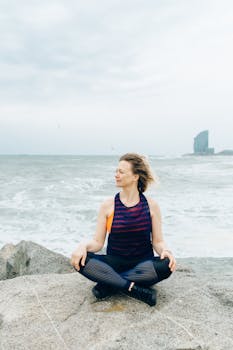 Woman in activewear meditating on a rocky beach with the sea in the background.