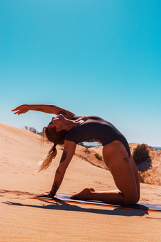A woman performs a yoga pose on a sunny beach dune, emphasizing flexibility and relaxation.