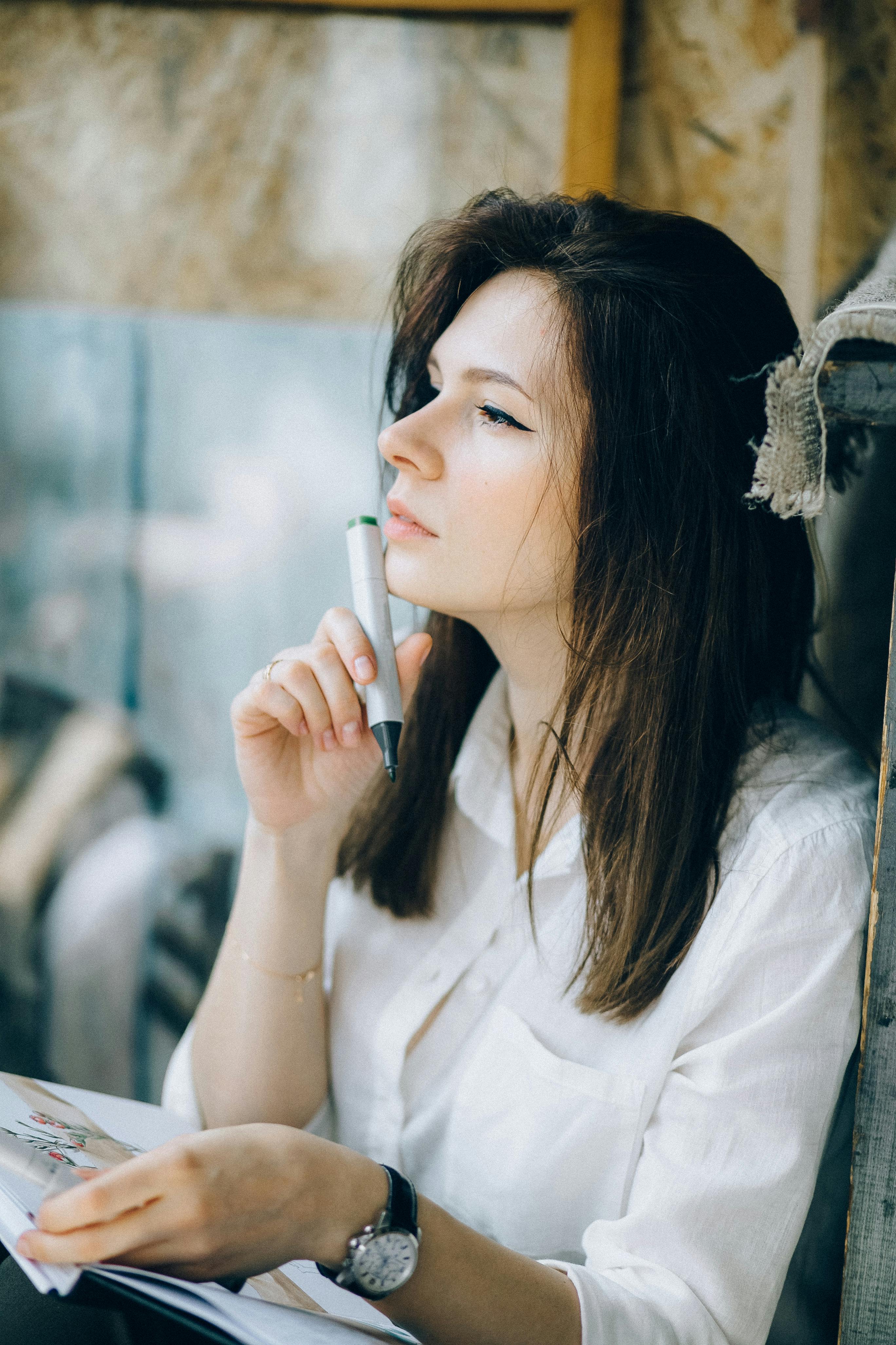 一位黑发女人在室内沉思，她穿着白色长袖，手里拿着笔