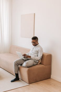 A man sits on a sofa, using a laptop in a minimalist home environment. Perfect shot for remote work themes.