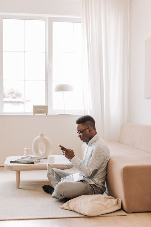 Free A man using a smartphone while sitting on the floor in a contemporary living room with neutral decor. Stock Photo