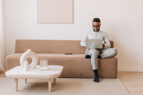 Free Adult man sitting on a sofa with laptop, focusing on work in a modern, minimalist room. Stock Photo
