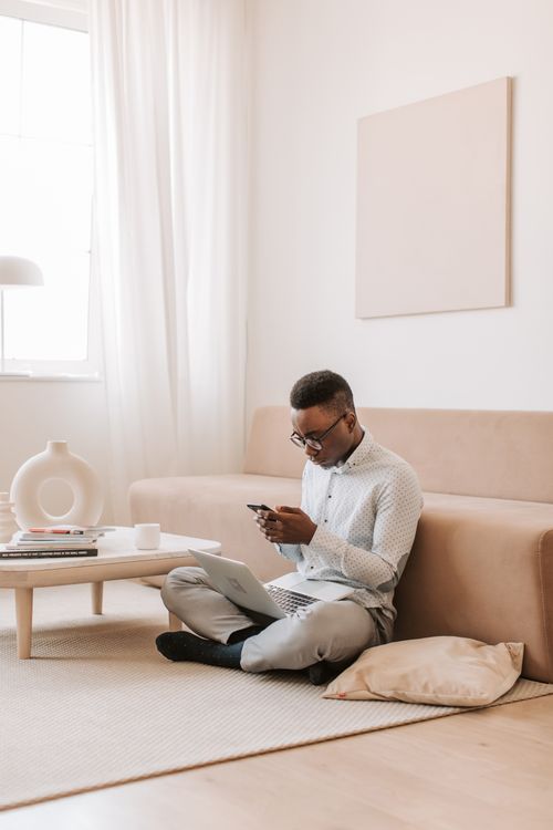 Free A man using a smartphone and laptop while sitting in a living room with a modern aesthetic. Stock Photo