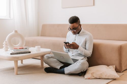 Free A man using a smartphone and laptop in a modern living room, highlighting wireless technology. Stock Photo
