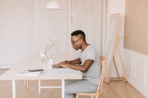 Free Young man in gray shirt working on a laptop indoors, embodying modern remote work lifestyle. Stock Photo