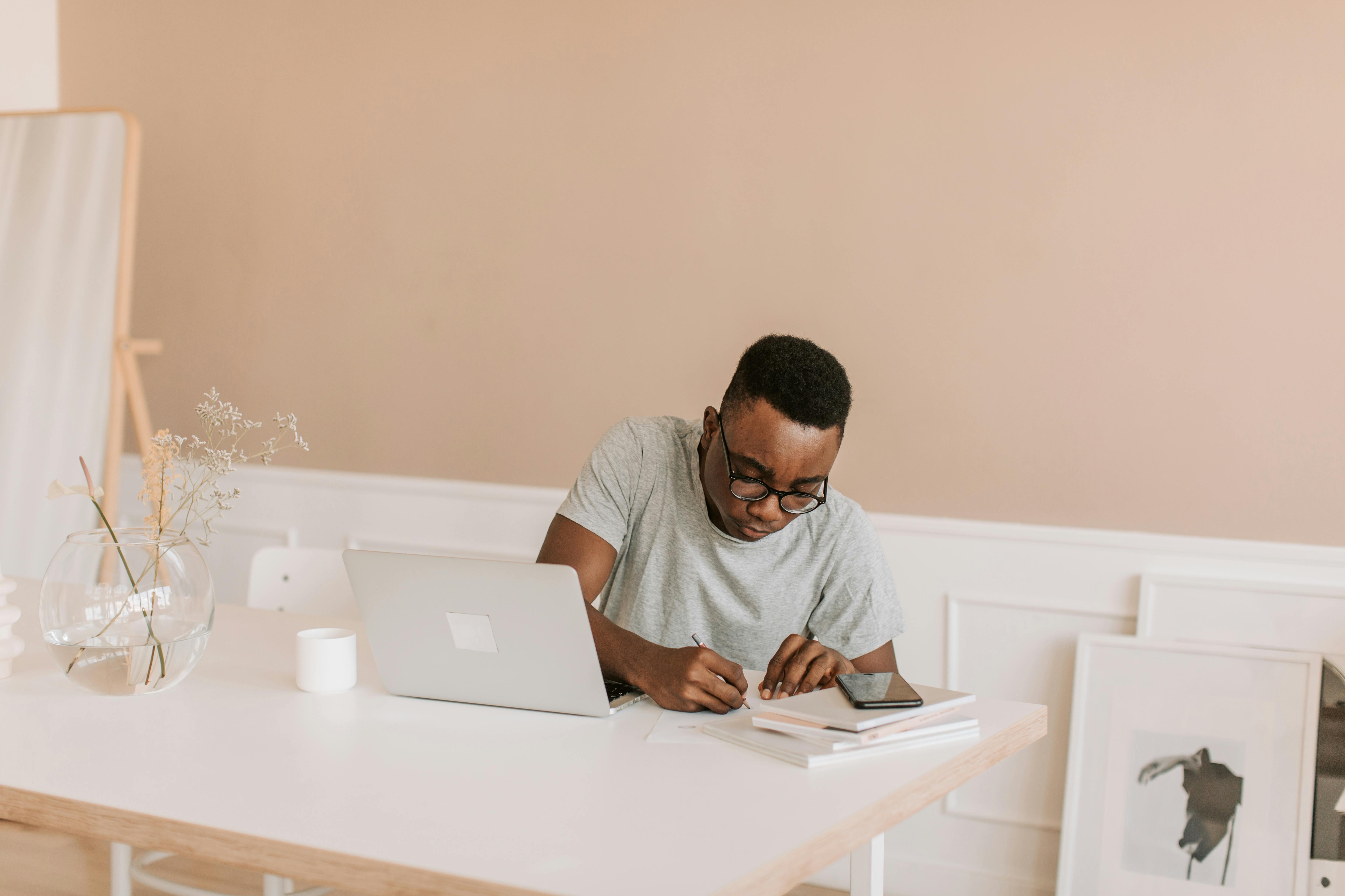 A man focuses on work at a minimalist home office, using a laptop and taking notes.
