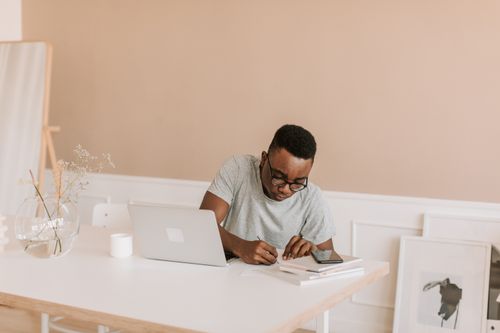 Free A man focuses on work at a minimalist home office, using a laptop and taking notes. Stock Photo