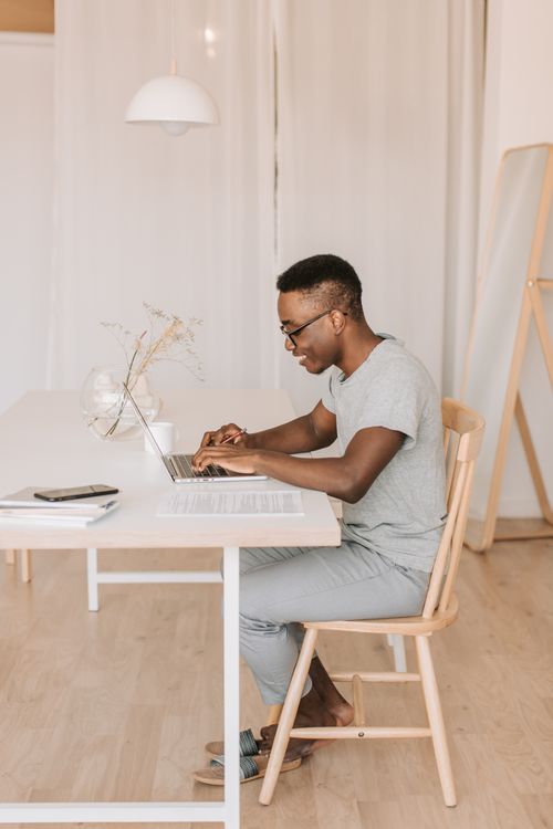 Free Side view of a smiling man working on a laptop at home. Minimalist decor with natural lighting. Stock Photo
