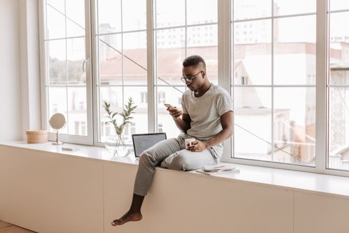 Free A man sitting on a window sill indoors, using a smartphone and holding a coffee cup. Stock Photo