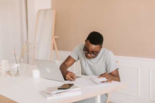 Free A man working from home on a laptop while taking notes and writing in a notebook. Stock Photo