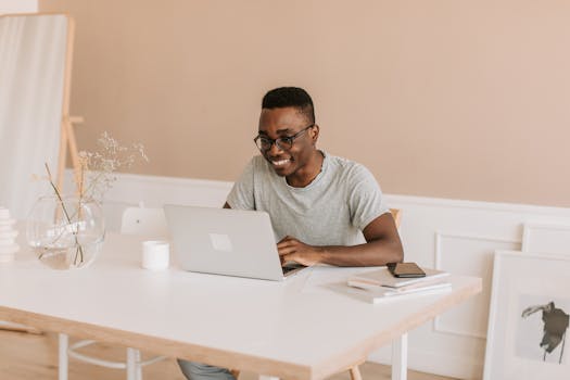 A man in gray shirt smiling while working on a laptop in a bright home office setting.
