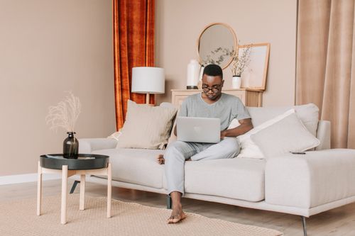 Free A man sits comfortably on a sofa using his laptop for remote work in a cozy living room. Stock Photo