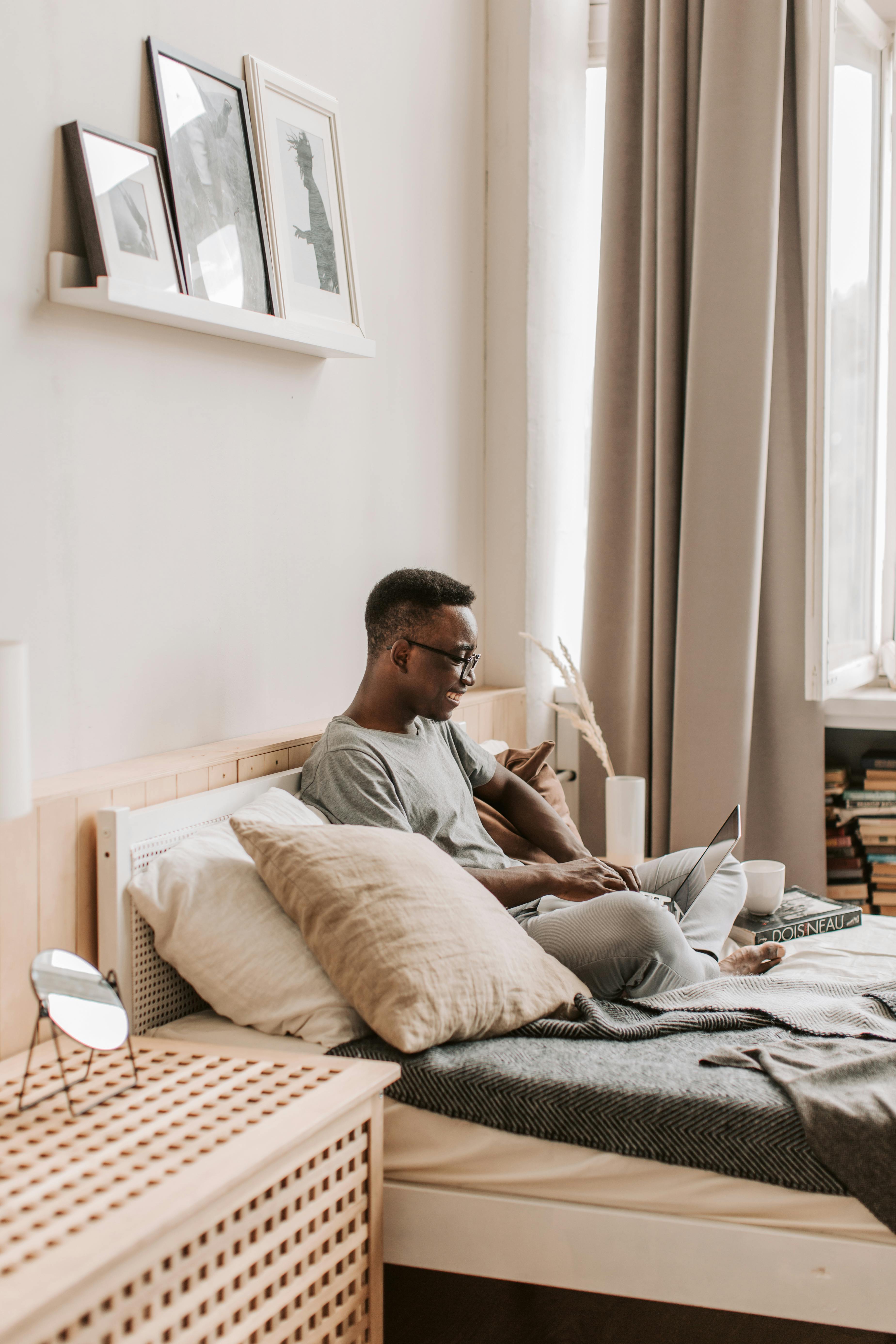 African American man using laptop for remote work, sitting on bed in stylish room.