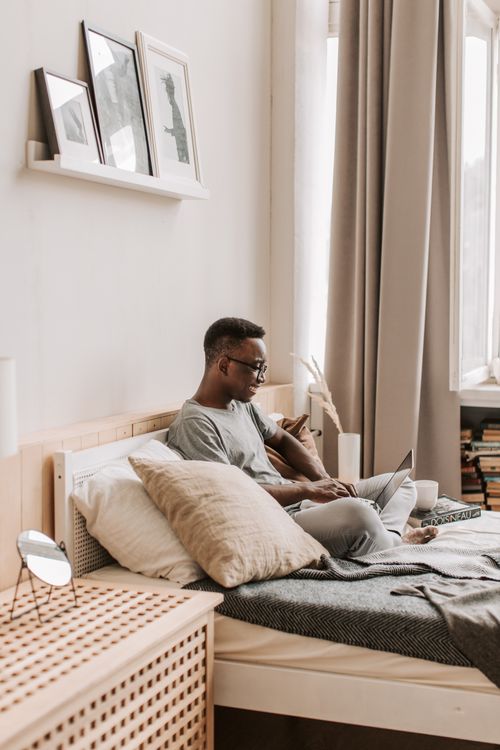 Free African American man using laptop for remote work, sitting on bed in stylish room. Stock Photo