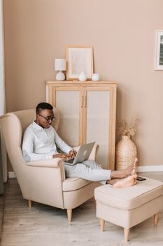 African American man comfortably working from home, sitting on a chair with a laptop.