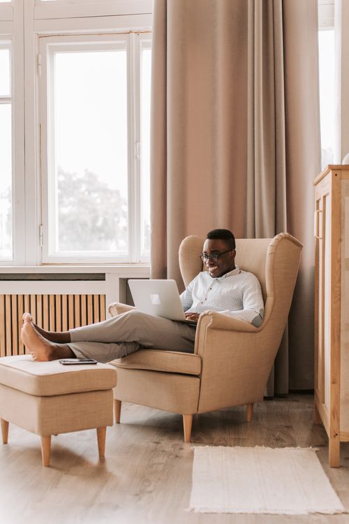 Free Man sitting in a comfortable chair, working remotely on a laptop in a bright, cozy home setting. Stock Photo