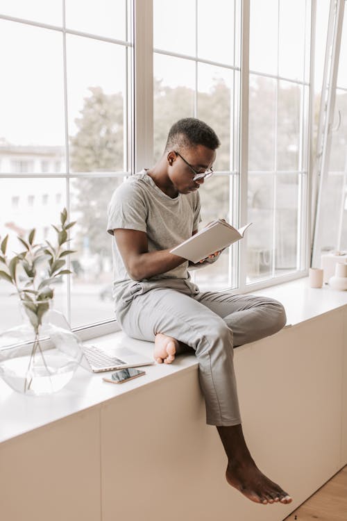 Free Man in Gray T-shirt and Gray Pants Sitting on White Table Using Silver Macbook Stock Photo