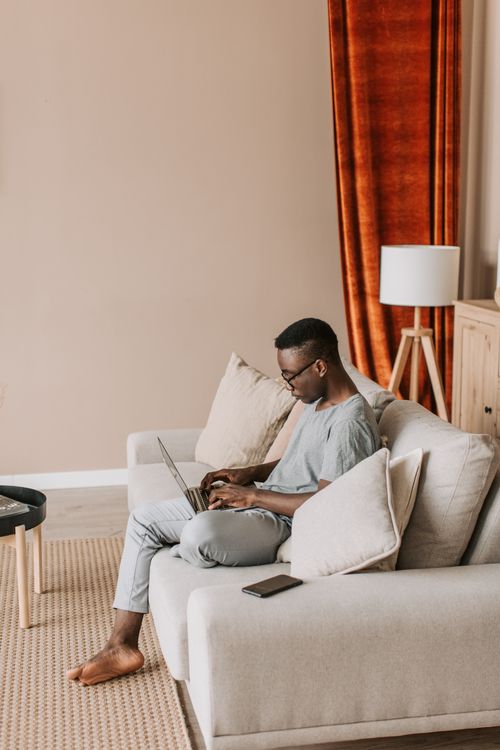 Free African American man using laptop on sofa for remote work in a cozy living room setting. Stock Photo