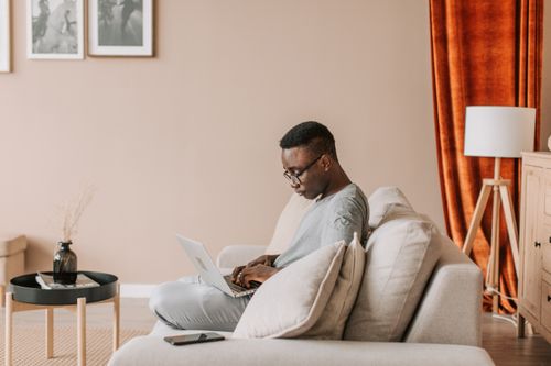 Free A man working remotely on a laptop while sitting comfortably on a sofa in a cozy living room. Stock Photo
