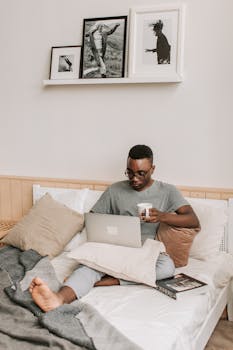 Relaxed man working on laptop in bedroom with coffee, embracing remote work lifestyle.