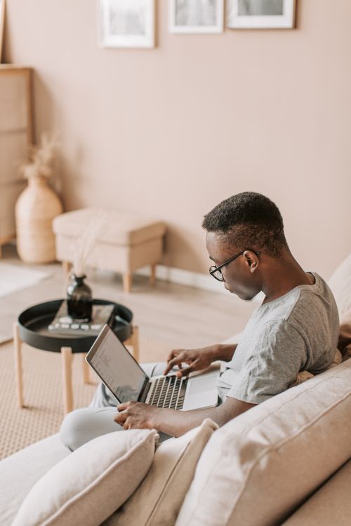 Free African American man working remotely on a laptop while sitting on a cozy sofa in a stylish living room. Stock Photo
