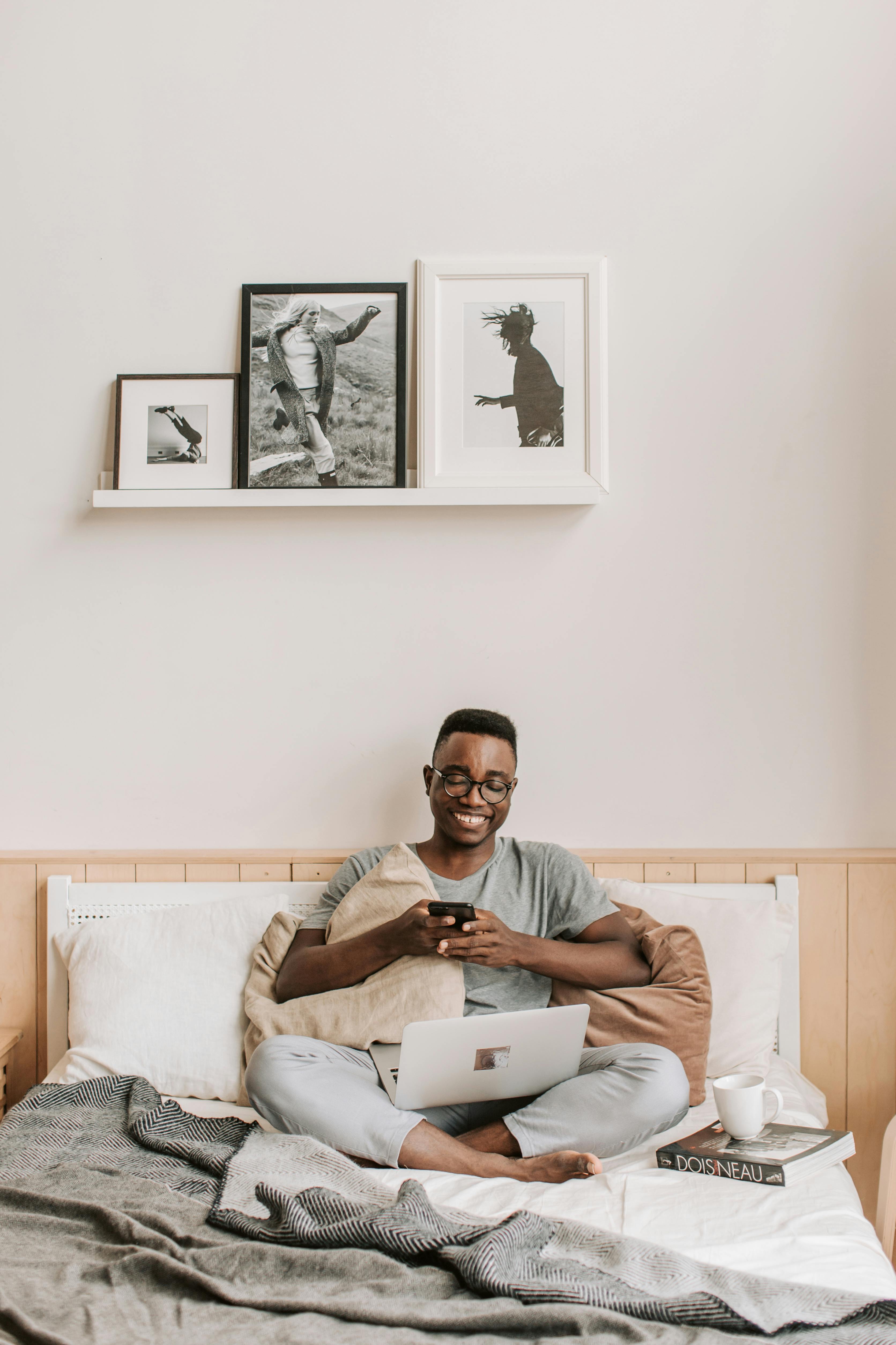 Smiling man sitting on bed texting on smartphone with open laptop.