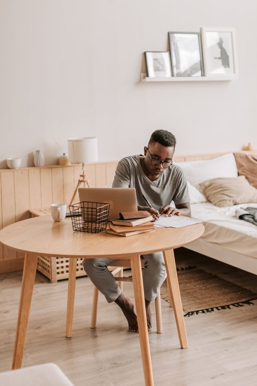 Free Man sitting barefoot at a table in a cozy bedroom, working on a laptop with notes. Stock Photo