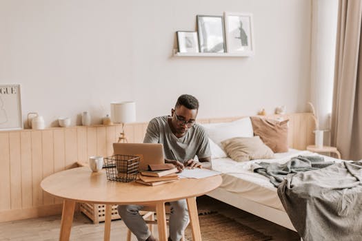 Mid-adult man works on his laptop in a cozy, well-lit bedroom, embodying the remote work lifestyle.
