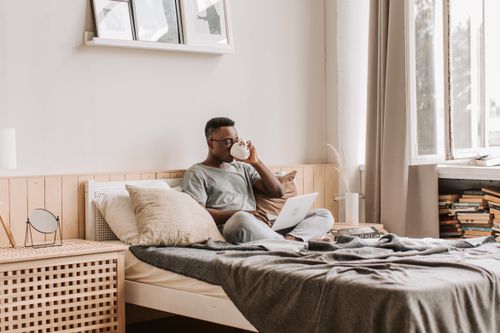 Free African American man sipping coffee while working on a laptop in a cozy bedroom setting. Stock Photo