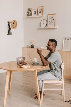 A man sits at a table using a smartphone and laptop in a stylish home office, smiling and relaxed.